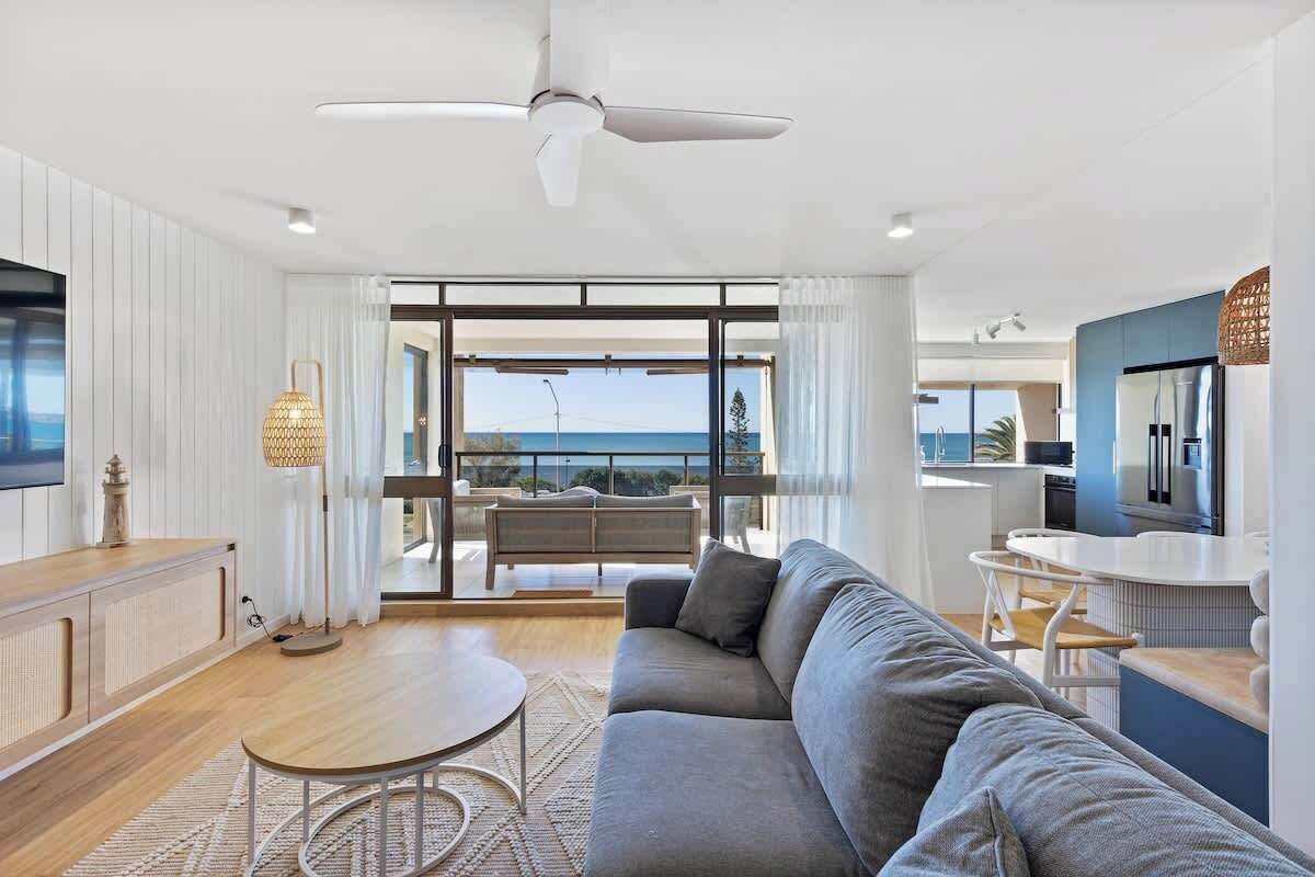 A modern living room with a sofa, a round coffee table, and a TV. There is a balcony with a view of the sea.