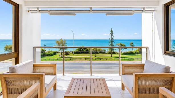 A balcony with a view of the ocean, wooden furniture, and a clear sky.