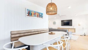 A modern dining area with a white table, wicker chairs, a bench, a hanging wicker light, a television, and a wall-mounted clock.