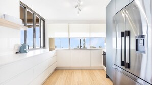 A modern kitchen with a stainless steel refrigerator, white cabinetry, a wooden floor, and a window with a view of trees and a clear sky.