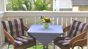 Ein Balkon mit Korbstühlen und einem Tisch, eine Vase mit gelben Blumen und Blick auf Bäume und ein Gebäude.