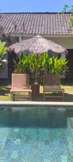 A pool area with two wicker chairs under a thatched umbrella, surrounded by lush greenery and a tiled-roof building. - Masaya Retreat Lombok (Kuta)