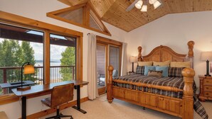 A bedroom with a wooden bed, a desk, and a chair, a balcony with a view of the lake, and a ceiling fan. - Luxury Lakefront Retreat on Lake Granby Near Snowmobiling (Grand Lake)