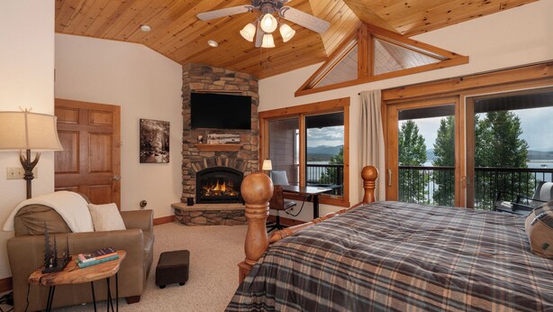 A bedroom with a stone fireplace, a bed, a desk, and a view of a lake. - Luxury Lakefront Retreat on Lake Granby Near Snowmobiling (Grand Lake)