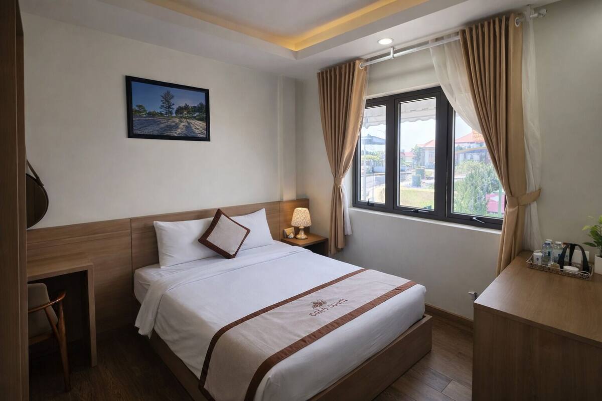 A hotel room with a bed, a desk, a chair, a lamp, a window with curtains, and a picture on the wall.