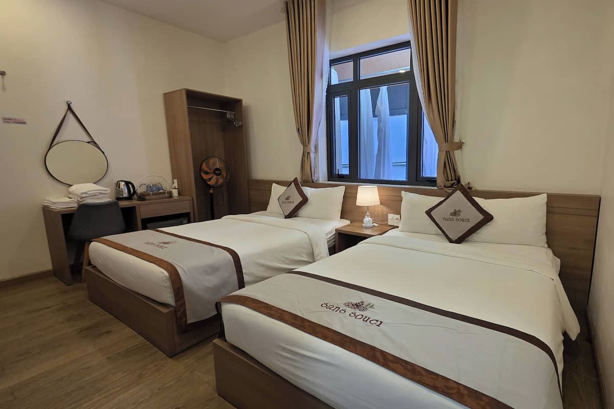 A hotel room with two beds, a desk with a chair, a fan, a window with curtains, and a sign on the wall.