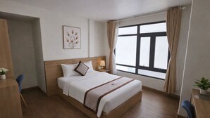 A hotel room with a bed, a desk, a chair, a nightstand, a lamp, a window with curtains, and a wall-mounted picture. - Vô Tư Hotel (Xuan Huong - Da Lat)