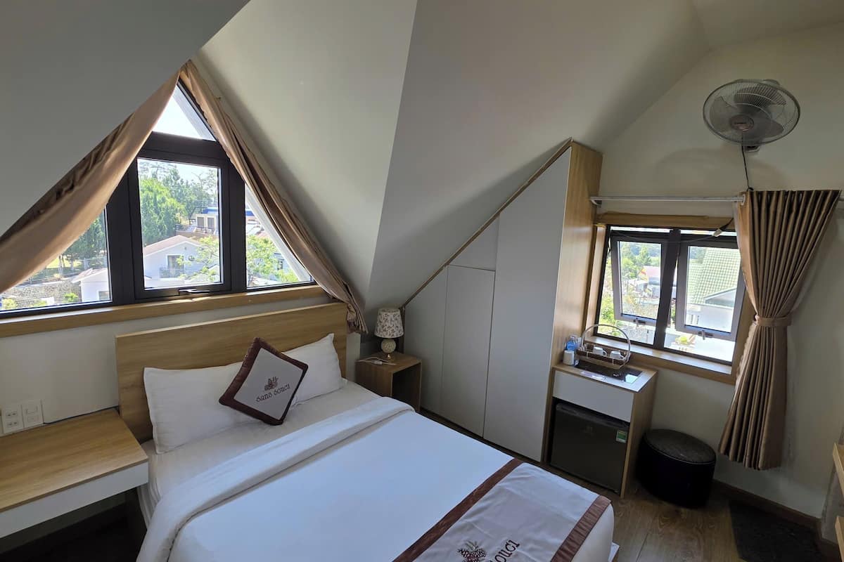 A bedroom with a bed, a desk, a chair, a fan, and a window with curtains.