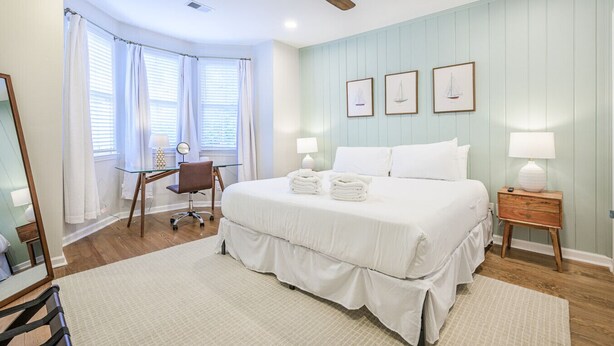 A bedroom with a large bed, a desk, a chair, a ceiling fan, and a window with curtains.