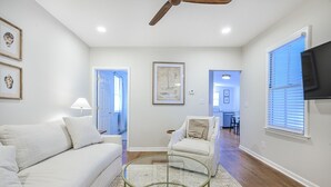 A spacious living room with a white sofa, a glass coffee table, and a white armchair. There is a ceiling fan, a framed map on the wall, and a television mounted on the wall.