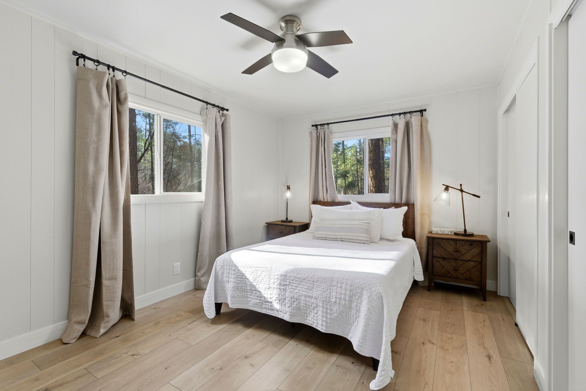 Un dormitorio con una cama, dos mesitas de noche, un ventilador de techo y una ventana con cortinas.