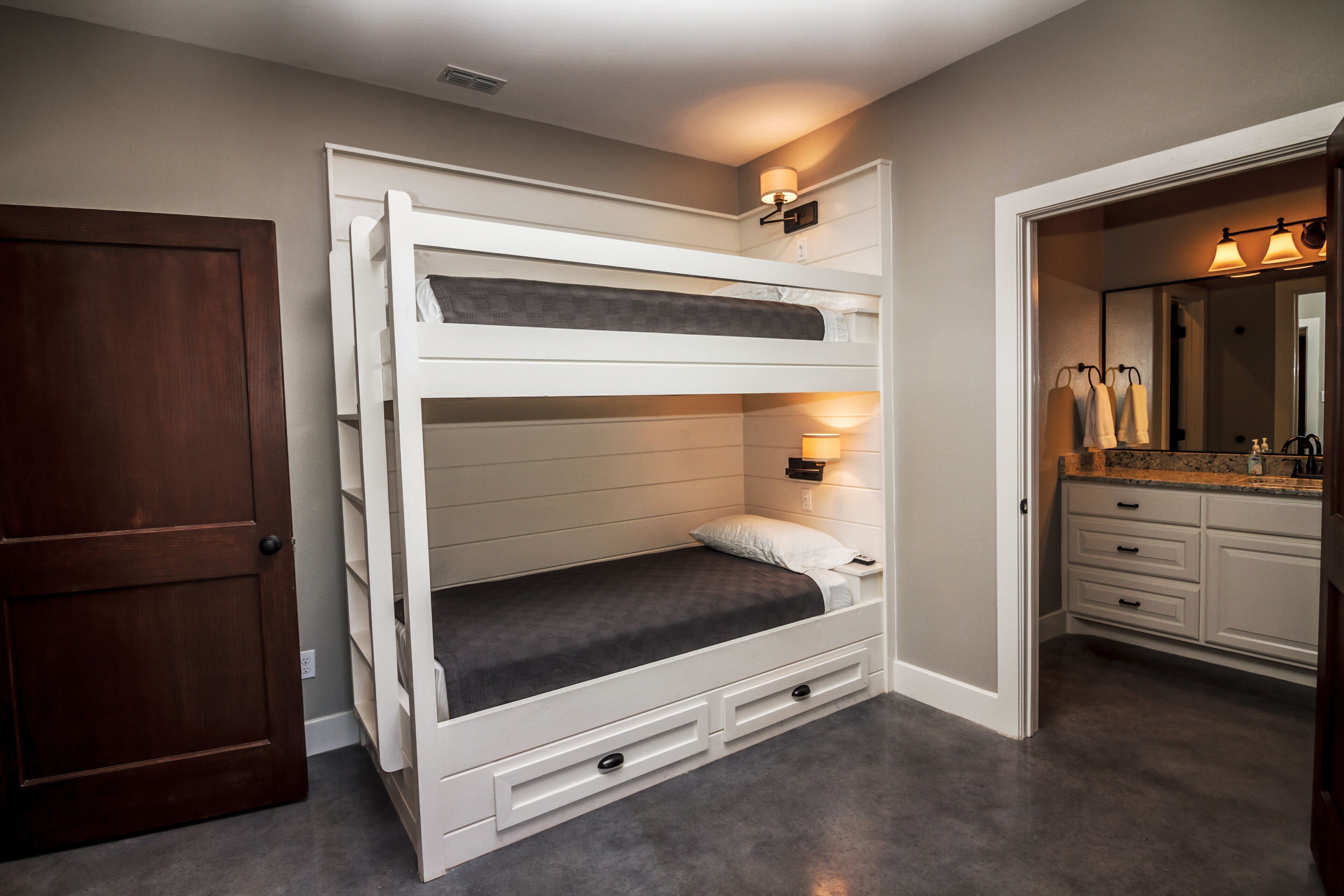 A bunk bed with a desk and drawers, a bathroom with a vanity and mirror, and a wooden door.
