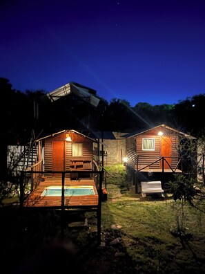 Two wooden cabins with outdoor decks and a hot tub, surrounded by trees and a dark sky.