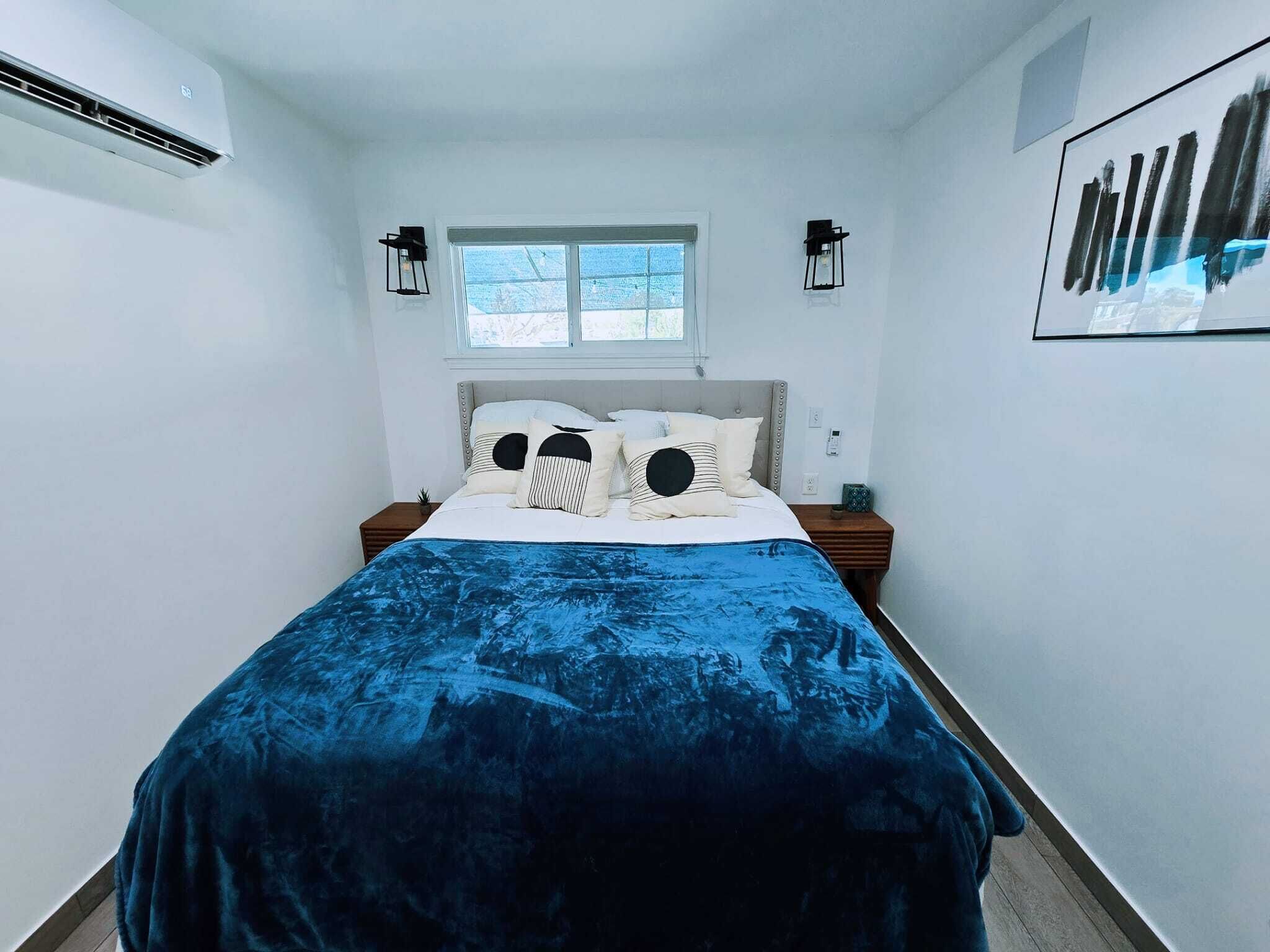 A bedroom with a bed, two bedside tables, a window, and a wall-mounted air conditioner.