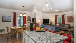 Una cocina moderna con encimeras de granito, un comedor con sillas de madera, una sala de estar con sofá gris y un televisor montado en la pared.