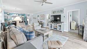Ein geräumiges Wohnzimmer mit Sofa, Couchtisch und Fernseher. Man sieht einen Deckenventilator, eine Küchenzeile und einen Esstisch.