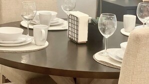 En spiseplads med et rundt bord dækket til fire personer, hvide tallerkener, kopper og vinglas. Der er en hvid orchid vase på bordet, og en lille potteplante ved siden af.