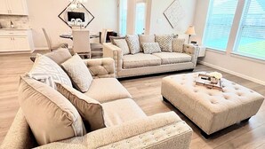 En rummelig stue med en hjørnesofa, skammel og et spiseområde med en lysekrone.