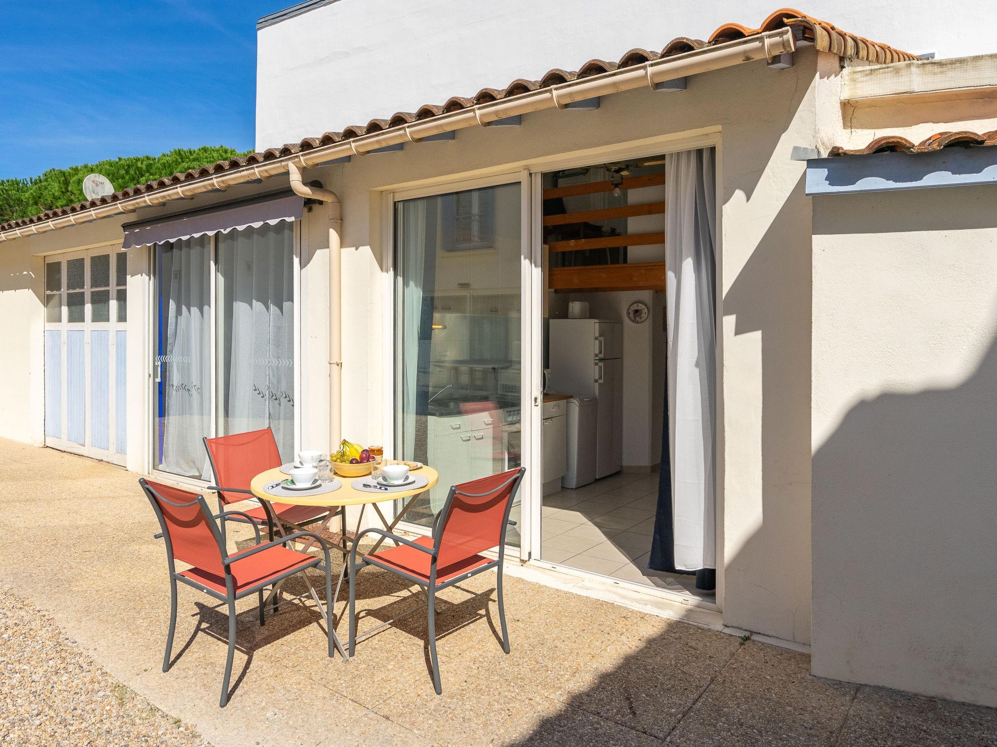 Un petit espace extérieur aménagé pour prendre les repas, avec une table et quatre chaises, une porte-fenêtre coulissante donnant sur une cuisine, et un bâtiment blanc doté d’un toit recouvert de tuiles.