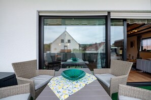 A balcony with wicker furniture, a table with a green bowl, and a view of a building through large glass doors.
