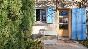 Uma casa pequena com porta azul e janelas, uma cadeira branca na parte externa e um arbusto verde em primeiro plano.