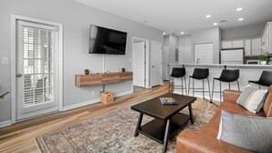 A modern living room with a sofa, coffee table, and TV. There is a kitchen area with bar stools and a dining table.