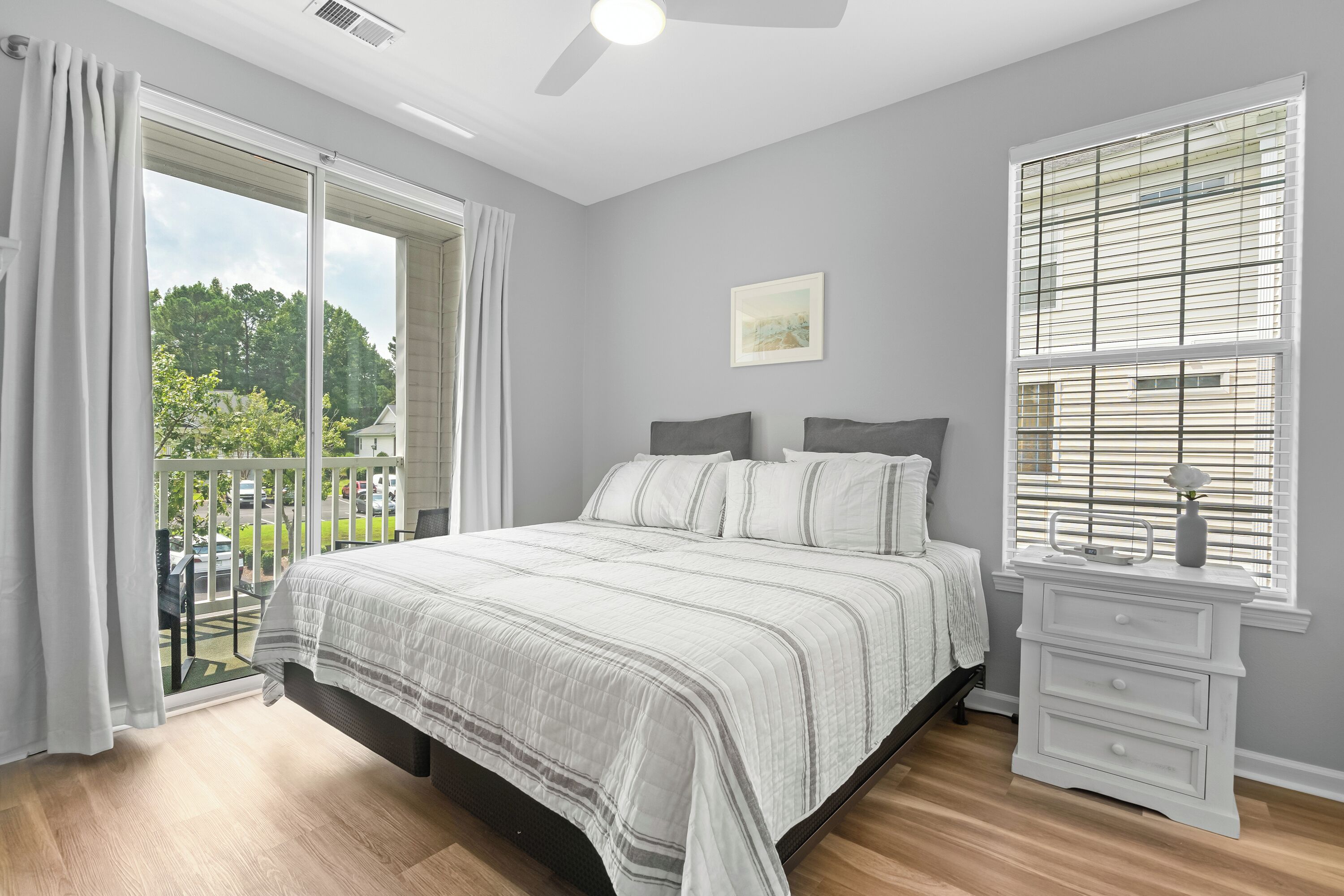 A bedroom with a large bed, a nightstand, a window with blinds, and a view of greenery outside.