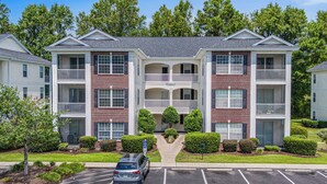 A multi-story apartment building with a central entrance, balconies, and a parking lot.