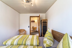 A bedroom with a bed, pillows, a chair, a wardrobe, and a door leading to another room.