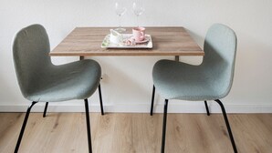Een eettafel met twee stoelen, een dienblad met een roze beker en een witte karaf, en twee lege wijnglazen aan de muur.