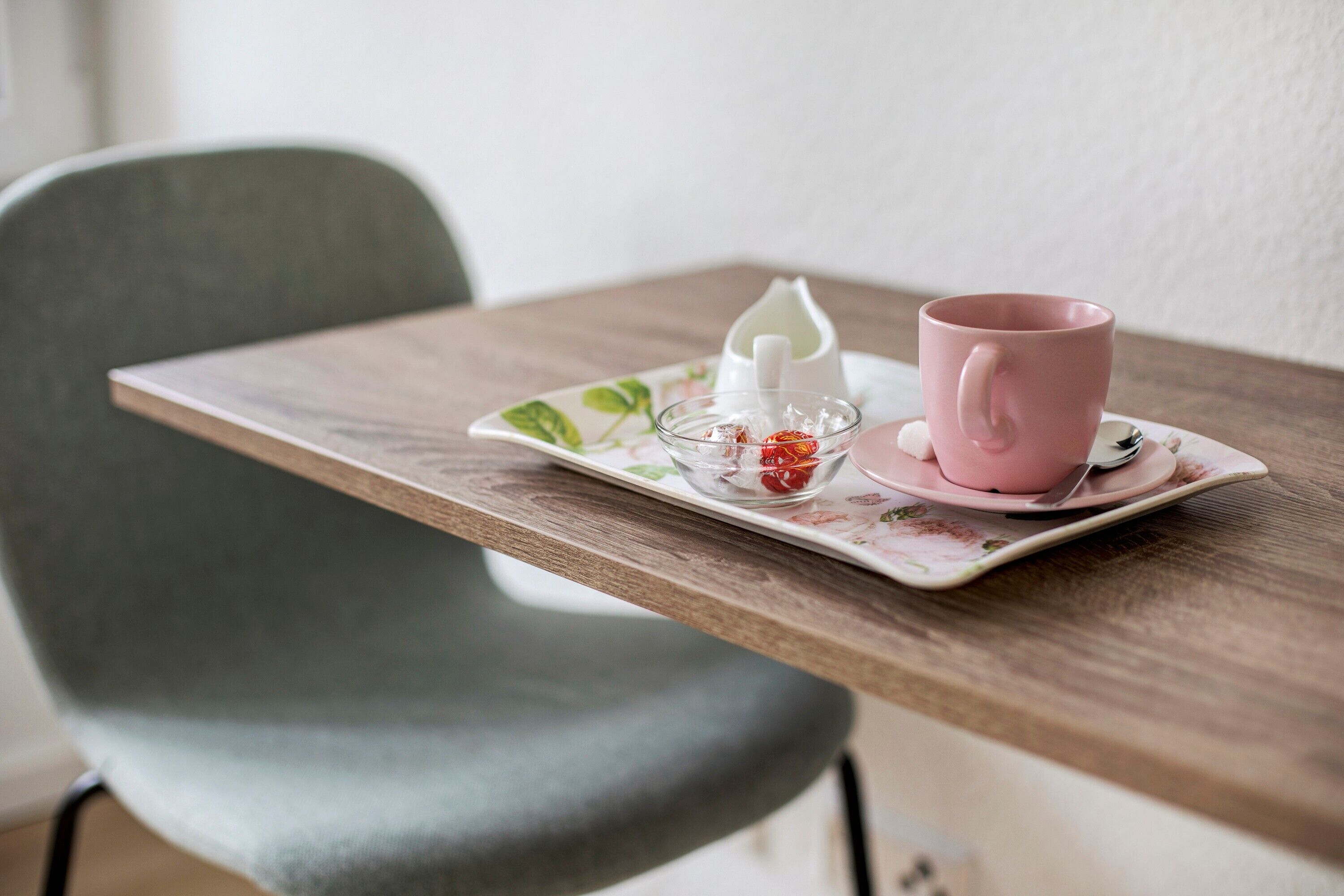 Una mesa con una taza y platillo rosa, un pequeño recipiente de vidrio y una jarra blanca sobre una bandeja estampada con motivos florales.