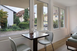 Une pièce comprenant une table à manger, deux chaises, un lit et de grandes fenêtres donnant sur le quartier.