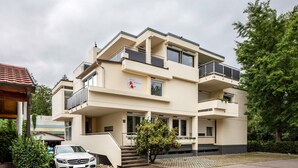 Un immeuble résidentiel à plusieurs étages, doté de balcons, avec une voiture garée devant et un petit arbre dans l’allée.