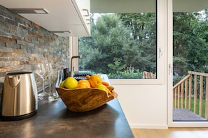 A kitchen with a stone backsplash, a wooden bowl with fruits, a kettle, and wine glasses.