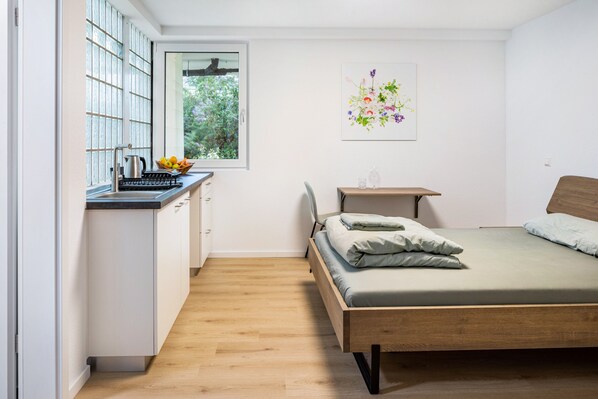 A bedroom with a wooden bed, a small bedside table, a window with a view of greenery, and a kitchen area with a kettle and fruit bowl.