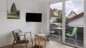 Un salón moderno con televisor de pantalla plana, un sofá gris, una mesita de madera con una copa de vino y un balcón con mobiliario exterior.