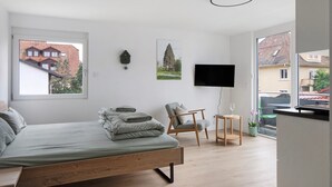 Un dormitorio con cama, una silla, un televisor y ventana con vista a edificios.