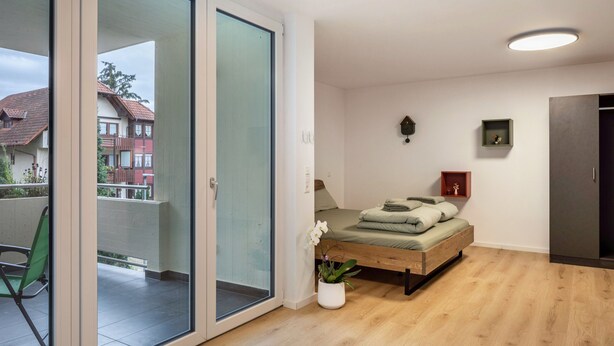 A modern bedroom with a sliding glass door leading to a balcony, a bed with a wooden frame, a small bedside table, and a potted plant. - Studio Apartment 'Belchen' with balcony and Wi-Fi (Bad Krozingen)