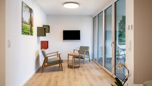 A modern living room with a flat-screen TV, wooden furniture, a small table, and a large window with a view. - Studio Apartment 'Belchen' with balcony and Wi-Fi (Bad Krozingen)