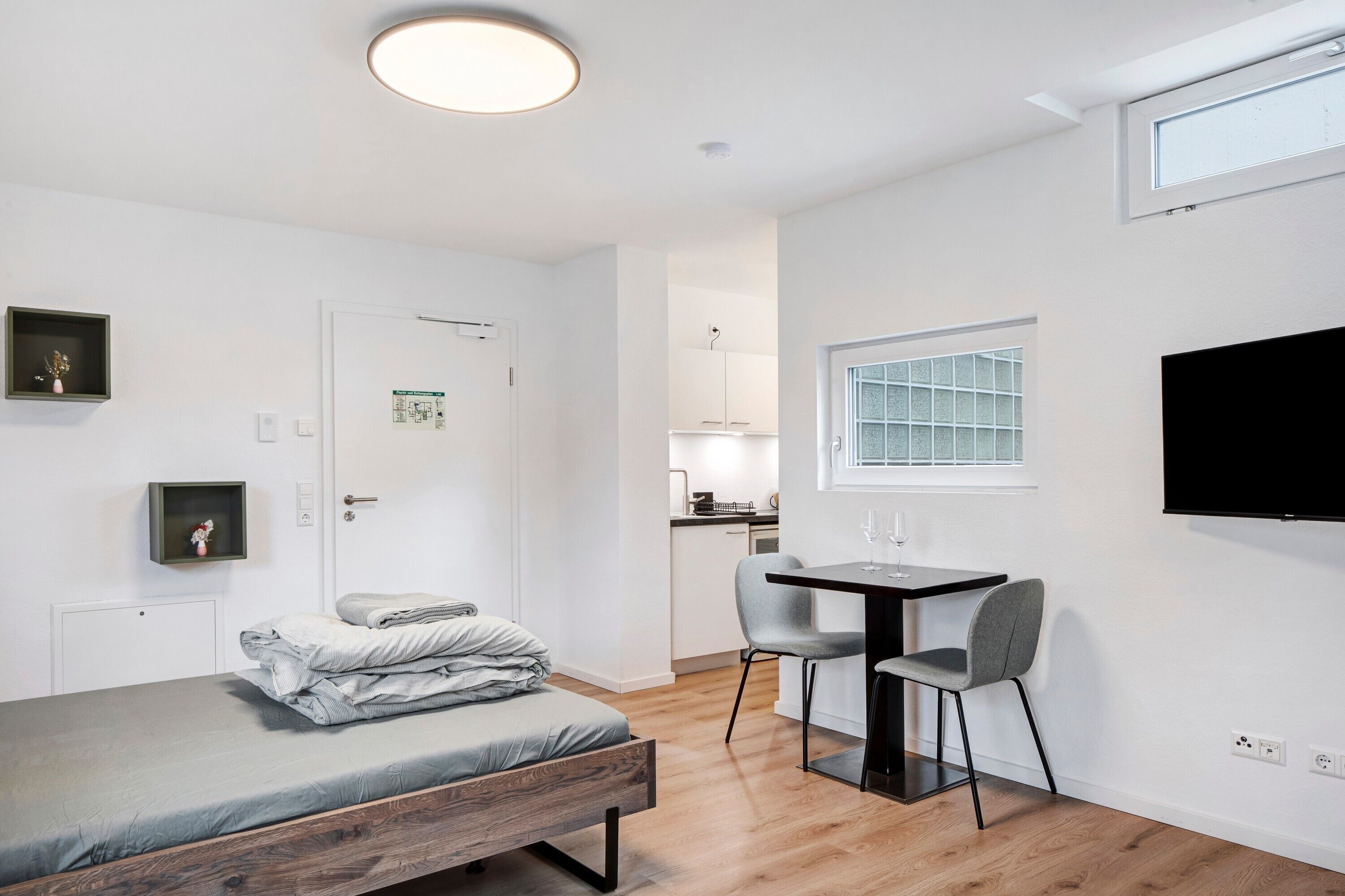 A compact room with a bed, a small table with two chairs, a TV mounted on the wall, and a window with a grid pattern.