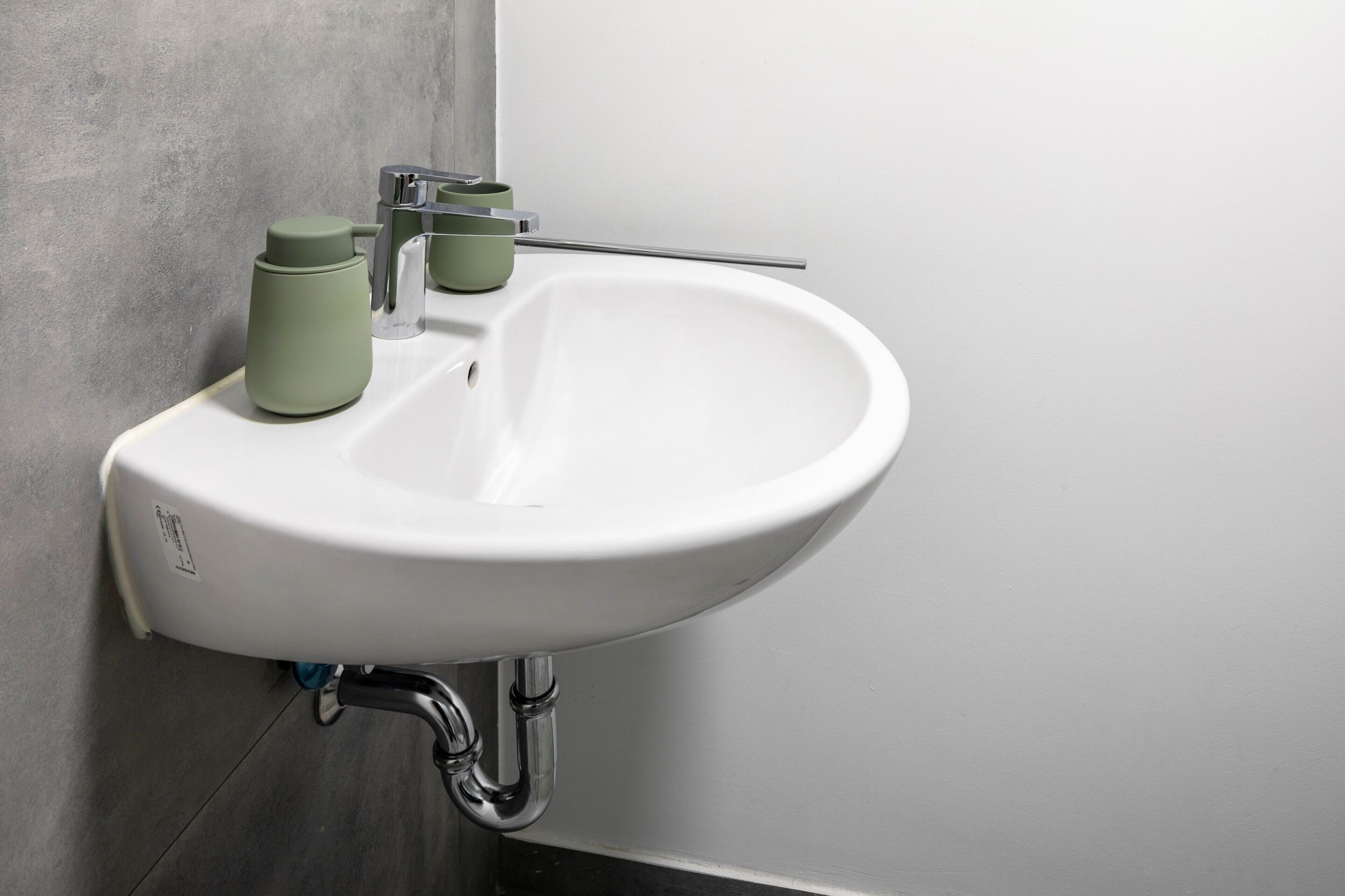 Ein weißes Waschbecken mit chromfarbenem Wasserhahn, eine grüne Tasse und ein Glas auf einer Arbeitsplatte.