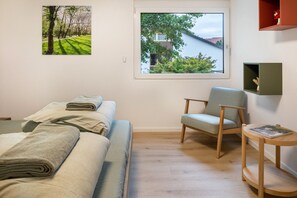 Ein Schlafzimmer mit Bett, Sessel, kleinem Tisch und Fenster mit Blick auf Bäume und ein Gebäude.