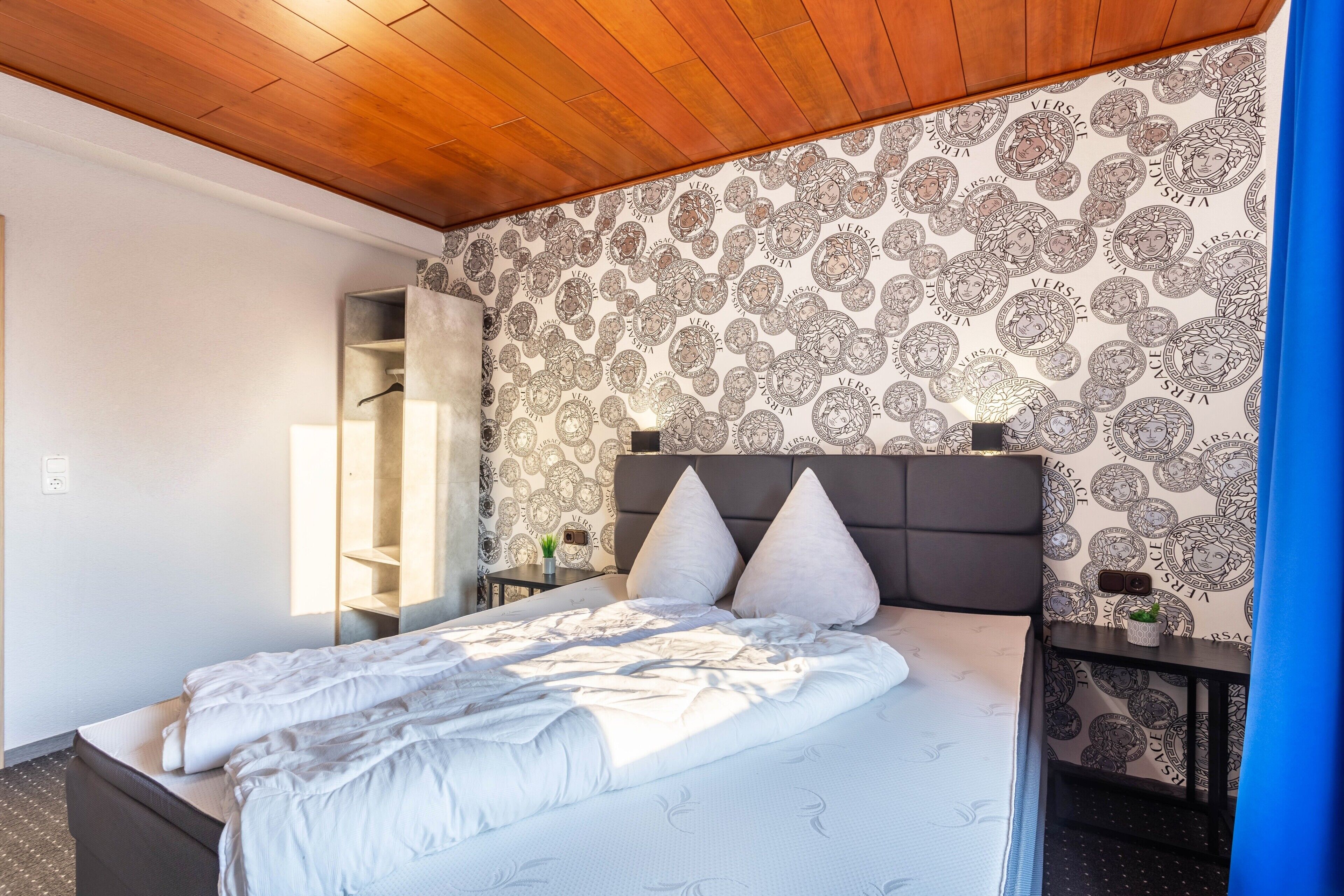 A hotel room with a bed, a nightstand, a mirror, and a patterned wallpaper.
