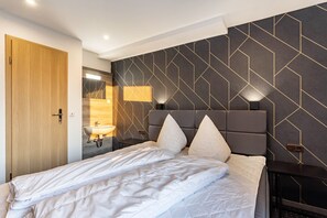 A modern hotel room with a large bed, a wooden door, a bathroom with a sink and mirror, and a geometric-patterned wall.