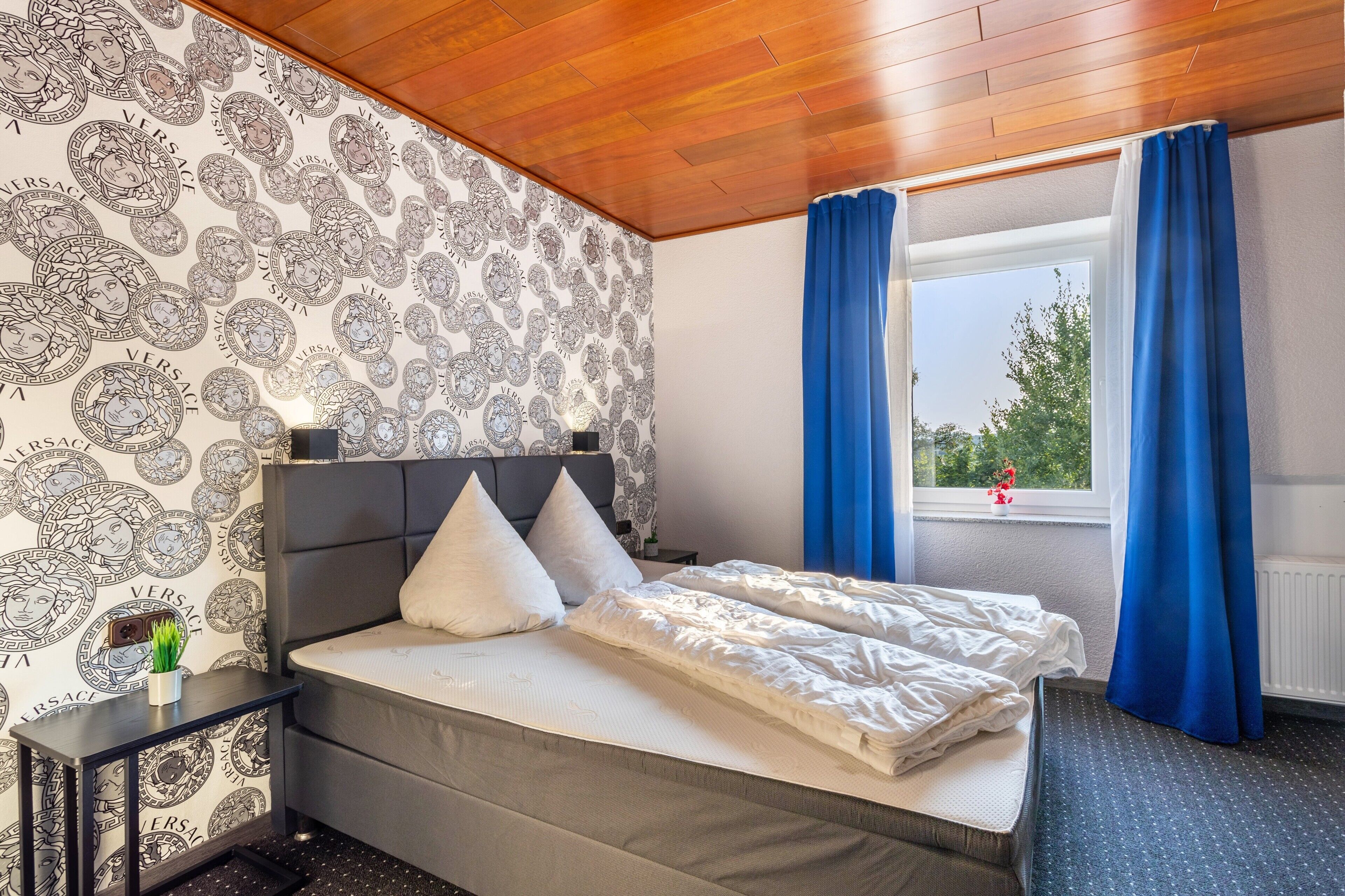 Ein Schlafzimmer mit einem Bett, einem Nachttisch, einem Fenster mit Vorhängen und einem Wandpapier mit Muster.