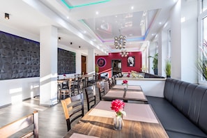 Ein modernes Restaurantinterieur mit hohen Decken, geometrischen Wandmustern und einem zentralen Kronleuchter. Tische sind mit dunklen Holzstühlen und schwarzen Sitzkissen angeordnet.