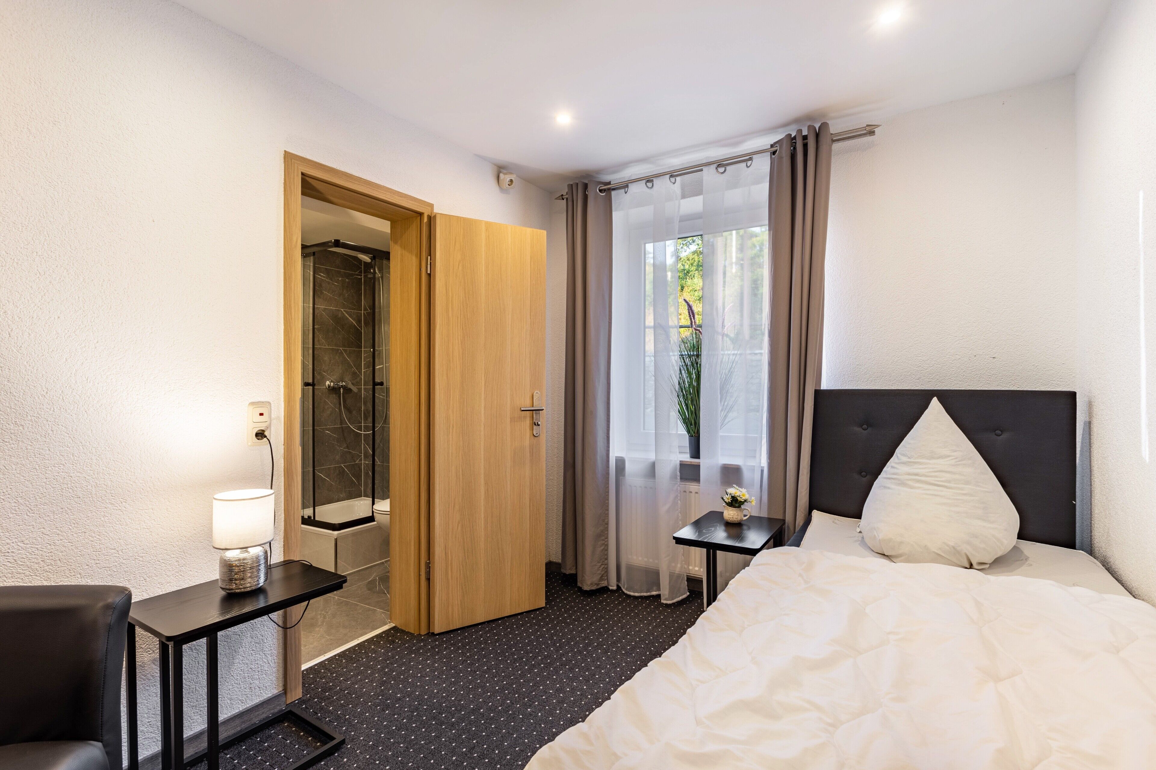 A hotel room with a bed, a bedside table with a lamp, a small table with a vase, a shower area, and a view of greenery outside.