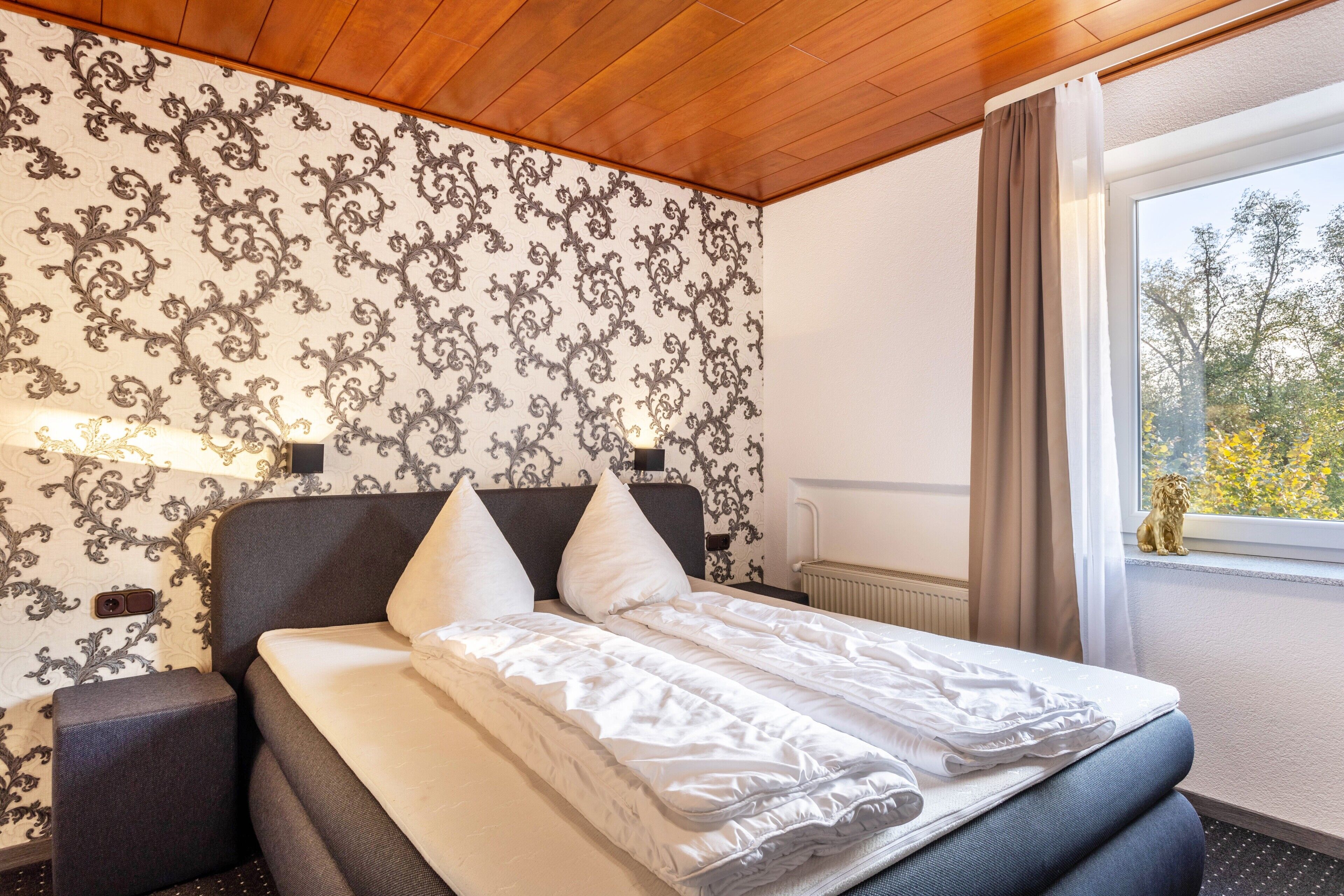Habitación de hotel con cama, mesita de noche, papel tapiz estampado y ventana con vista a árboles.
