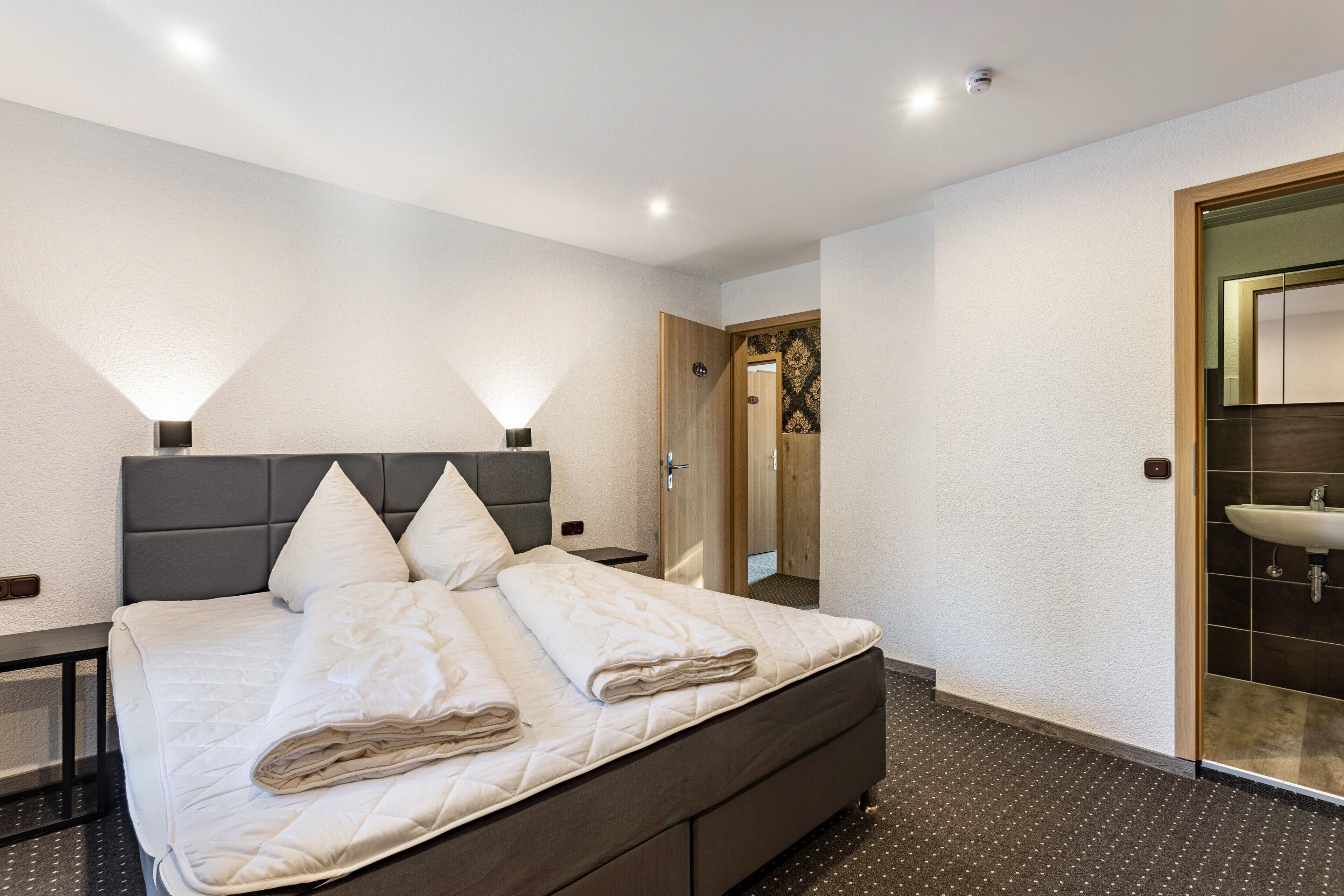 Una habitación de hotel moderna con una cama grande, mesitas de noche, un espejo y un baño con lavamanos.