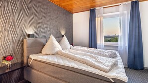 Een slaapkamer met een bed, nachtkastje, lamp, raam met gordijnen en uitzicht op het omliggende landschap.